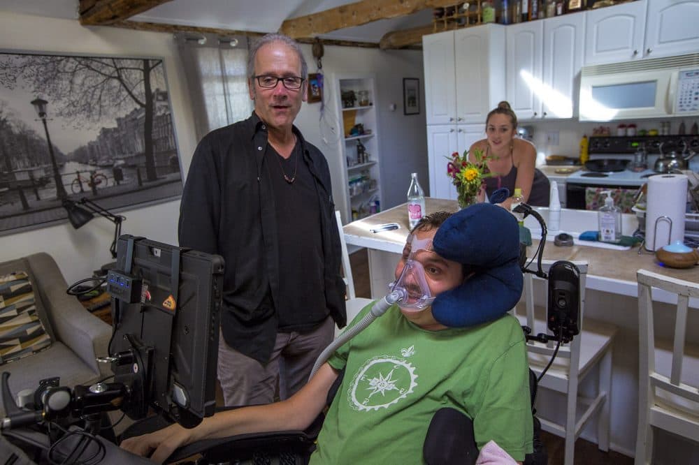 In this 2016 photo, Bobby Forster communicates with Hoffman with a computer that uses an eye-tracking device mounted onto his wheelchair. Forster's wife, Casey, is behind them. Casey Forster and Hoffman had just discussed the prospect of a family road trip to New Orleans. (Jesse Costa/WBUR)