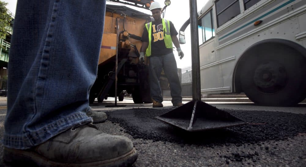A proposed tax on incomes over $1 million will fund Massachusetts schools and infrastructure. Unless, of course, it won't.
Pictured: Boston Public Works workers Victor Duret, feet only at left, and Tyrone Odom, behind, both of Boston, fill a pothole with asphalt in Boston's Charlestown neighborhood. (Steven Senne/AP)
