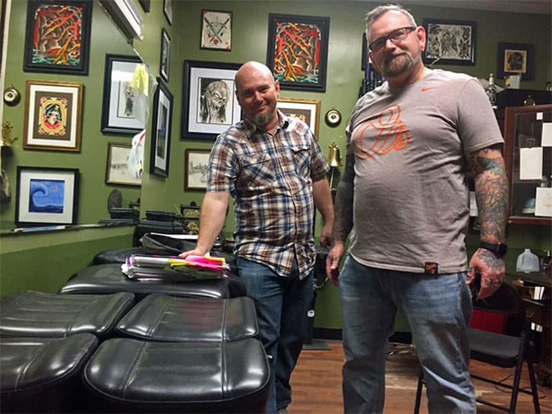 Dave Ente and Dave Cutlip in Southside Tattoo stand next to a pile of files on the tattoo cover-ups they've been working on over the past year. (Mary Rose Madden for WBUR)