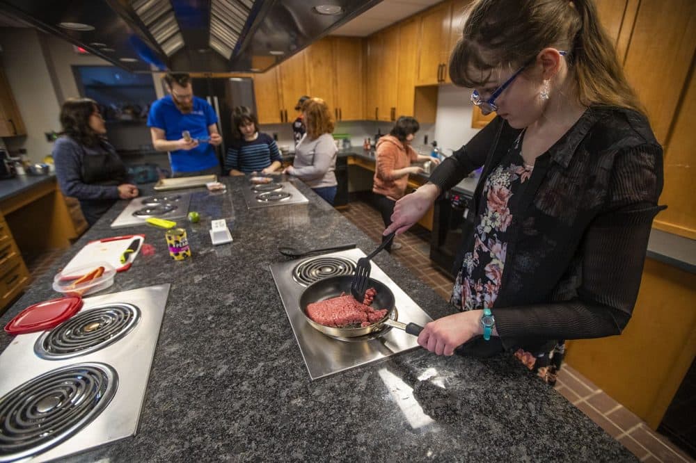 Taylar Bogdanovic cooks ground beef on the stovetop for English muffin pizzas. The kitchen is bustling with activity. (Jesse Costa/WBUR)