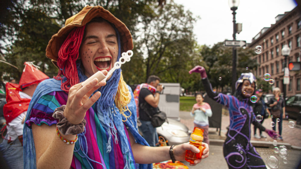 Sivan Spector, dressed as the “anti-productivity clown,” blows bubbles while on roller skates, as another artist dances in the background. (OJ Slaughter for WBUR)
