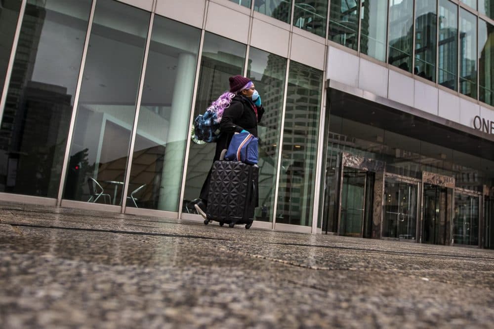A woman with a face mask traveling with a suitcase walks toward South Station by One Financial Center. (Jesse Costa/WBUR)