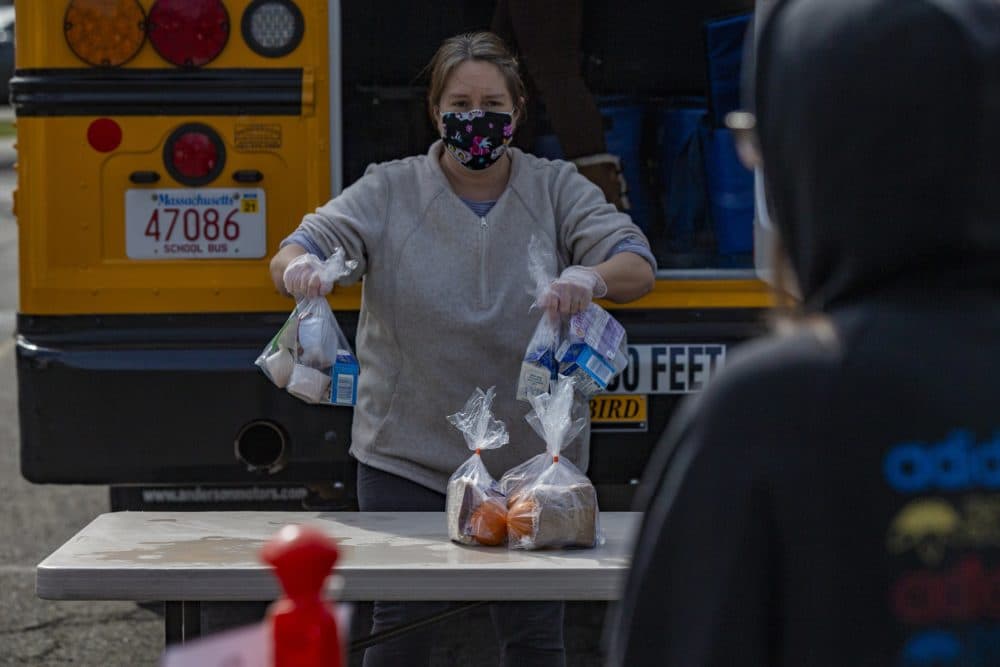 Salem public schools bus driver Sharon Moulton places bagged breakfast and lunch meals onto a table while a student waits ten feet away at pilon designated as a safe distance. Salem Public Schools is delivering "grab-and-go" style breakfast and lunch to all children under the age of 18 at designated areas around the city. (Jesse Costa/WBUR)