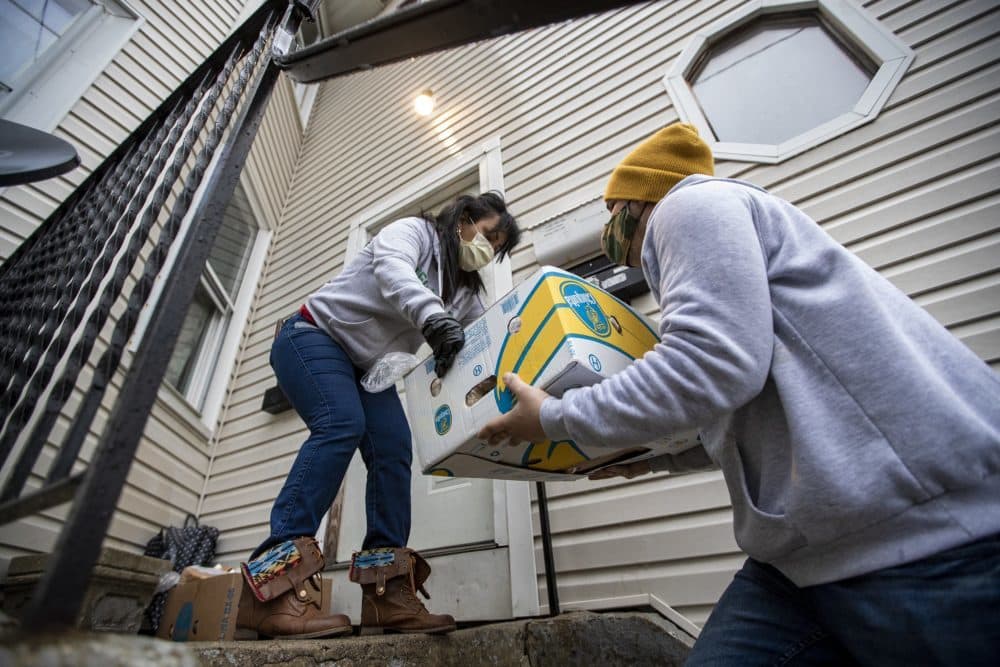 Reymer Pineda entrega a Gladys Vega una caja de comida donada para dejarla frente a una casa. Todos los miembros de ese hogar han contraído COVID-19 y saldrán a recoger la caja una vez Gladys les diga que pueden salir. (Jesse Costa/WBUR)