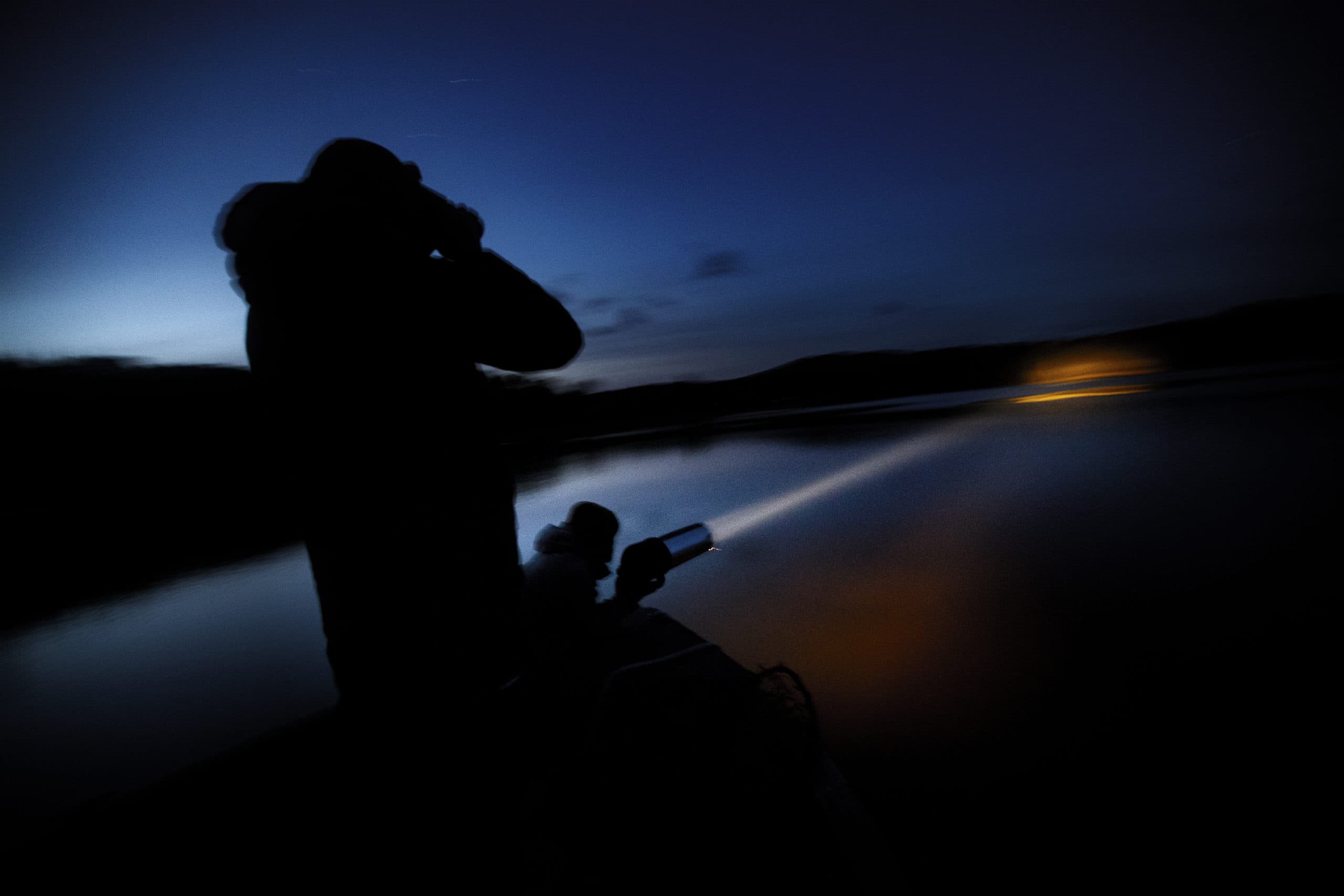 Mark Burton shines a spotlight across the water as Chris Persico uses a pair of binoculars to find loon chicks on Rangeley Lake in Maine. (Jesse Costa/WBUR)