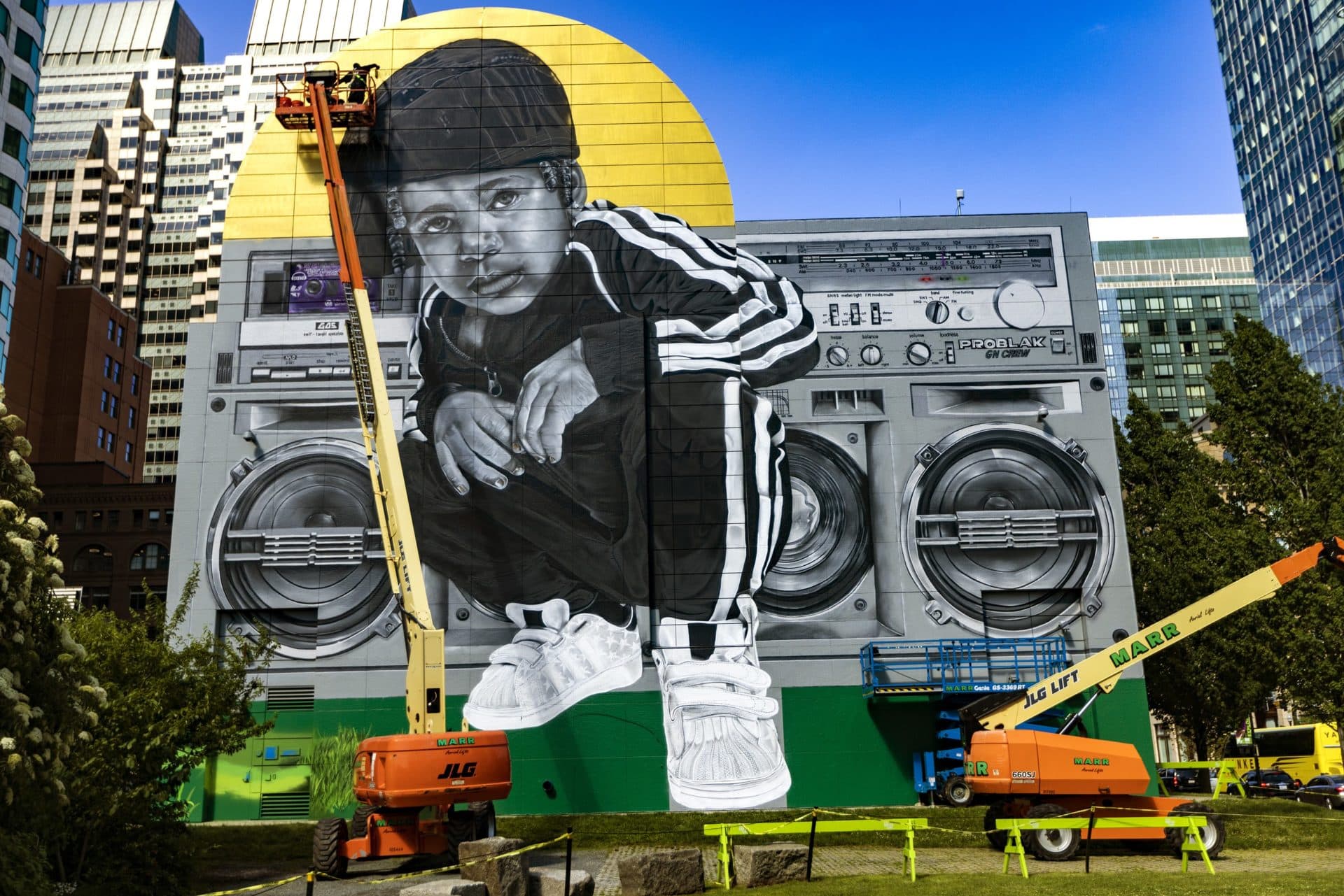 High above on a knuckle lift, Rob “ProblaK” Gibbs works on “Breathe Life Together,” the next mural to be featured on the façade of the Dewey Square Tunnel Air Intake Structure on the Rose Kennedy Greenway. (Jesse Costa/WBUR)