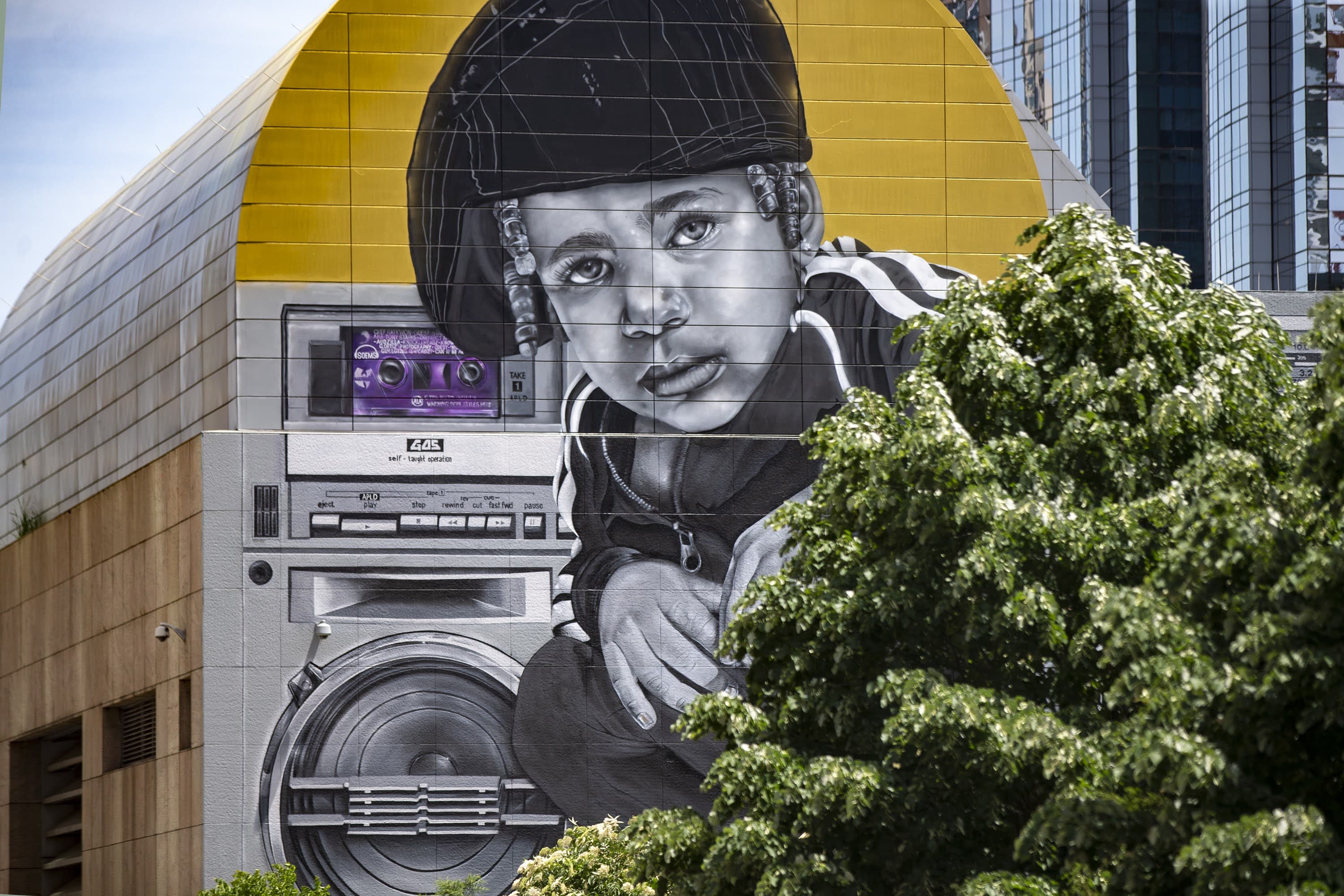 The likeness of the daughter of muralist Rob “ProBlak” Gibbs peers around the trees in Dewey Square in Gibbs’ work “Breathe Life Together,” the next mural to be featured on the façade of the Dewey Square Tunnel Air Intake Structure on the Rose Kennedy Greenway. (Jesse Costa/WBUR)