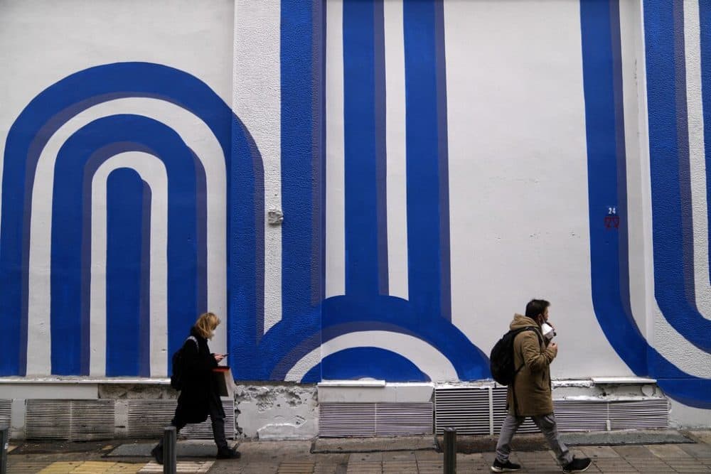 Pedestrians wearing face masks to curb the the spread of coronavirus, pass in front of the mural "Waves" by the artist Adioshpe in Athens, Greece, Wednesday, Jan. 12, 2022. According to health authorities Tuesday, the surge of the Omicon variant in Greece seems to have "slowed down." (AP Photo/Thanassis Stavrakis)